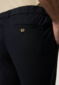 Marina blå byxor med elastisk midja och en enkel knappdetalj på bakfickan. Tillverkade av ett slätt textilmaterial.