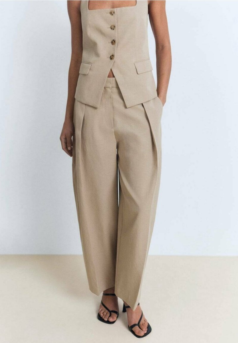 Beige op maat gemaakte vest met vierkante halslijn en vier knopen, gecombineerd met wijde broek met plooien, aangevuld met zwarte sandalen.