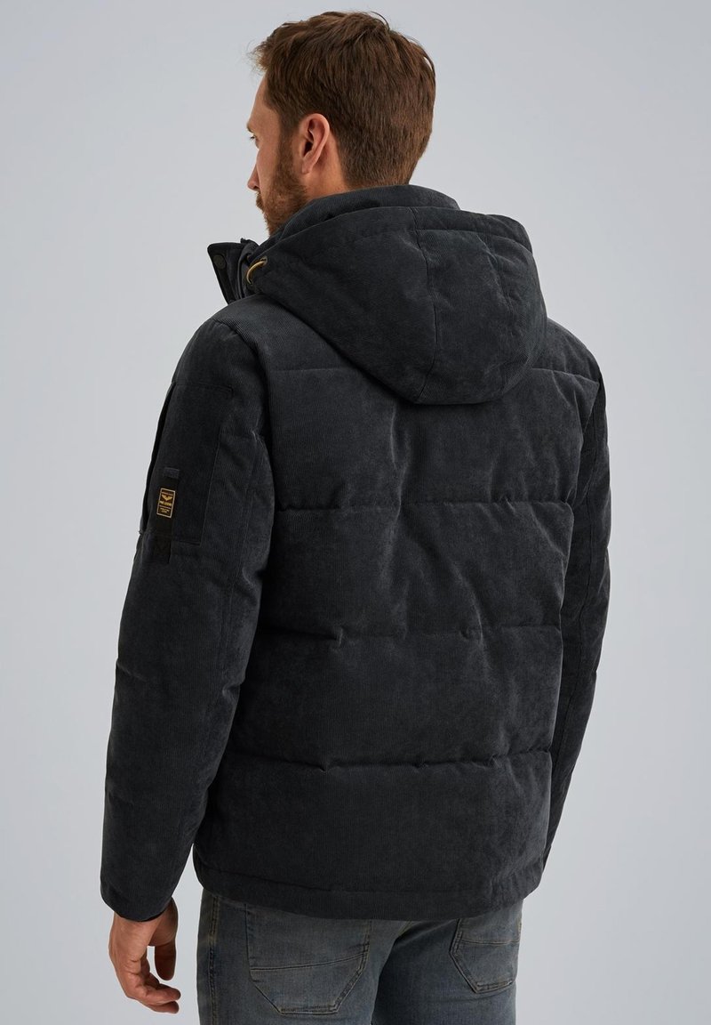 Chaqueta de pana negra acolchada con capucha, que presenta una superficie texturizada y sutiles detalles de costura. Incluye un parche con el logo en la manga izquierda.