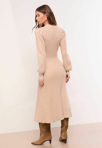Könnyű bézs színű kötött midi ruha bordázott textúrával, puffos ujjakkal és testhez simuló derékrésszel. Barna velúrból készült térdig érő csizmával kombinálva.