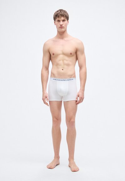 White cotton boxer briefs with elastic waistband featuring "POLO RALPH LAUREN" logo in navy. Model stands barefoot, facing forward.