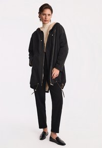 Czarna parkowa kurtka z kapturem, złotym zamkiem błyskawicznym, ściąganym dołem i bocznymi kieszeniami. Nosi ją osoba w kremowym swetrze z dzianiny i czarnych spodniach z błyszczącymi mokasynami.