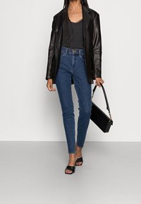 Mujer vestida con una chaqueta de cuero negra, camiseta negra, jeans ajustados azules y tacones abiertos negros, sosteniendo un bolso negro con cadena.