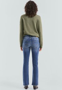 Groene gebreide trui gecombineerd met blauwe wijde jeans. De trui heeft een gestructureerde afwerking, terwijl de jeans achterzakken en lichte stiknaden hebben.