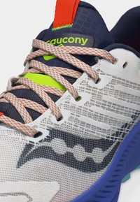 Κοντινό πλάνο ενός παπουτσιού τρεξίματος Saucony με λευκό δίχτυ, ναυτικό μπλε και νέον κίτρινα στοιχεία, πορτοκαλί και λευκά κορδόνια και μπλε σόλα.