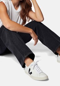 Personne portant un jean noir à jambes larges et des baskets blanches avec un logo noir, assise avec un bras reposant sur le genou sur fond blanc.