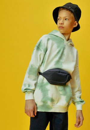 Hoodie in Weiß und grünem Batik, schwarze Gürteltasche, Jeans und schwarzer Bucket Hat vor einem gelben Hintergrund.