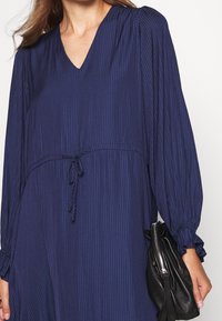 Námořnický modrý pruhovaný šaty s výstřihem do V, dlouhými nafouklými rukávy a staženým pasem na šňůrku. Obsahuje texturovanou látku a malou černou kabelku.