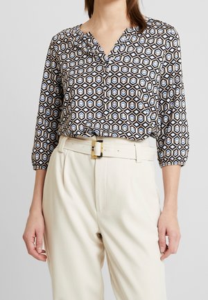 Femme portant un chemisier à manches longues à motifs, rentré dans un pantalon crème taille haute avec une ceinture assortie.