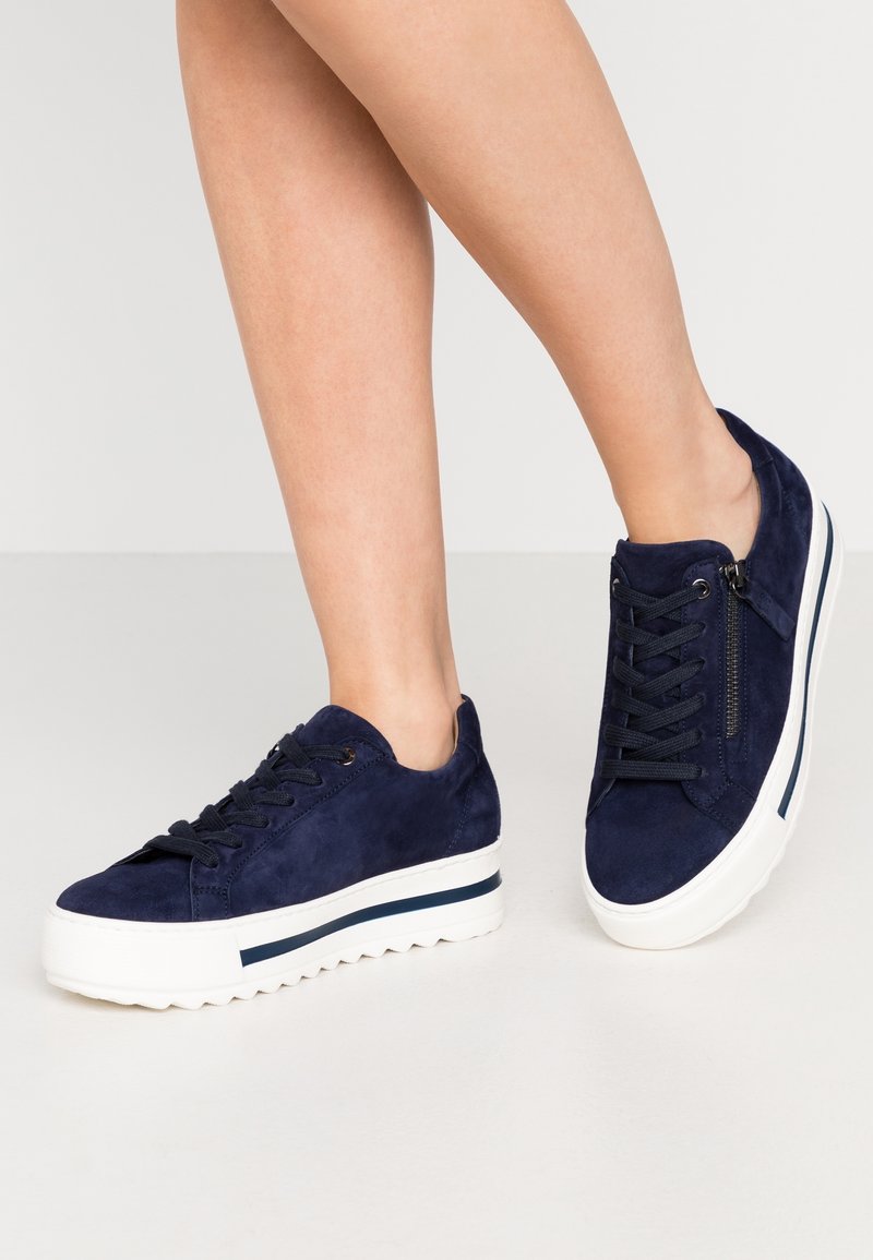 Jambes portant des baskets à plateforme en daim bleu marine avec des lacets noirs, une fermeture éclair sur le côté, et des semelles épaisses blanches avec une rayure bleu marine.