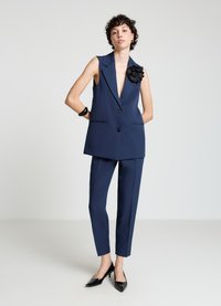 Blazer sans manches bleu marine avec col en V profond, pantalon slim, et accent floral noir. Des talons noirs brillants complètent l'ensemble.