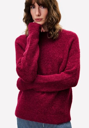 Vrouw met schouderlang golvend bruin haar, draagt een textuurrijke dieprode trui en blauw gestreepte broek, leunt met haar kin op haar hand.