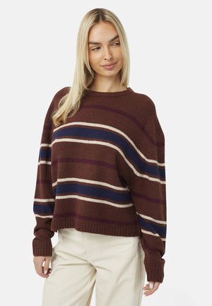 Vrouw met lang blond haar die een bruine trui draagt met marineblauwe, paarse en beige strepen en lichte broek, staand tegen een witte achtergrond.