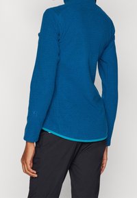 Modrá texturovaná bunda s vysokým límcem, s kontrastním spodním lemem v odstínu tyrkysové. Dlouhé rukávy s elastickými manžetami a diskrétním logem.