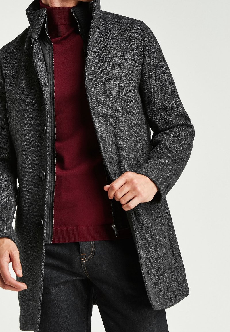 Homme portant un manteau en chevrons gris foncé par-dessus une couche intérieure noire zippée et un pull à col roulé bordeaux avec un jean foncé.