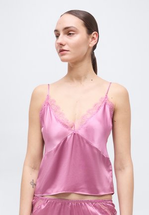 Jauna moteris tamsiais plaukais surištais uodegėlėje, vilkinti rožinę satininę palaidinę su nėrinių apdaila ir derančius šortus, žvelgianti į kairę.