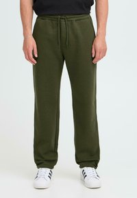Olivegrüne Jogginghose mit lockerer Passform, weichem Stoff, elastischem Bund und Kordelzug. Kombiniert mit weißen Turnschuhen mit schwarzen Streifen.