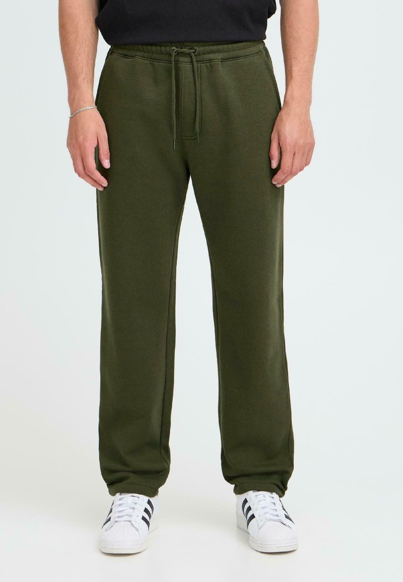 Olivegrüne Jogginghose mit lockerer Passform, weichem Stoff, elastischem Bund und Kordelzug. Kombiniert mit weißen Turnschuhen mit schwarzen Streifen.