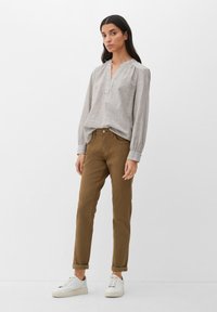 Jasnoszara bluzka z długim rękawem, z dekoltem w kształcie litery V i marszczonymi ramionami, w połączeniu z brązowymi spodniami zwężającymi się ku dołowi, z podwiniętymi mankietami, oraz białymi trampkami.