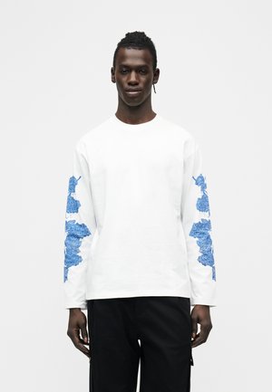 Mann trägt weißes langärmliges Hemd mit blauen Blumenmustern auf den Ärmeln und schwarze Hose, steht vor weißem Hintergrund.