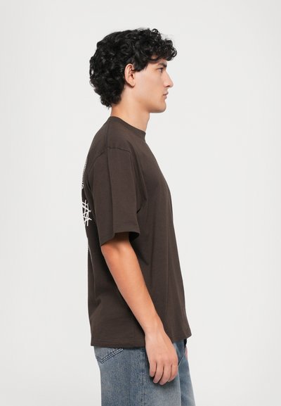 Ragazzo giovane con capelli ricci che indossa una maglietta oversize marrone scuro e jeans blu, rivolto a destra contro uno sfondo bianco semplice.