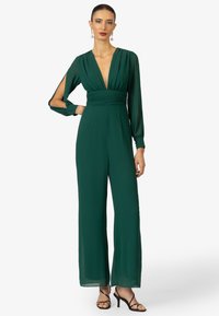 Groene jumpsuit met lange mouwen en een diepe halslijn, met geplooide taille. Zachte, vloeiende stof; brede broekspijpen, gedragen met hoge sandalen.
