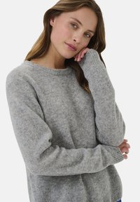 Šviesiaplaukė moteris su rudais plaukais, vilkinti pilką megztinį, atsirėmusi viena ranka į galvą, baltoje fone.