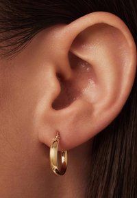 Boucle d'oreille en or, petite et lisse, présentée dans une oreille percée. La boucle d'oreille possède une surface brillante et une forme simple et arrondie.
