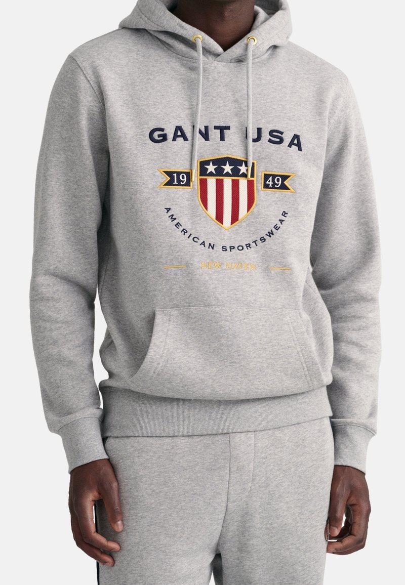 Homme portant un sweat à capuche gris clair et un pantalon de survêtement, le sweat à capuche arbore un écusson avec des étoiles et des rayures, texte "GANT USA 1949 American Sportswear".