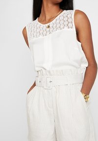 Top bianco senza maniche con scollo in pizzo, abbinato a pantaloni a vita alta a righe chiare con una ampia cintura e tasche frontali.