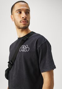 Camiseta negra de algodón con cuello redondo y mangas cortas, que presenta texto bordado en blanco en el pecho. Se lleva un bolso negro con correa ajustable cruzado en el cuerpo.