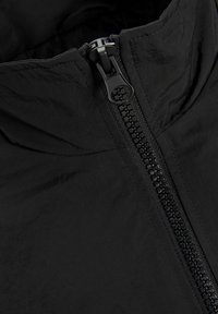 Veste noire en tissu lisse et texturé, avec une fermeture éclair sur toute la longueur et un col haut. La fermeture éclair présente une finition mate.