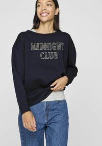 Sudadera de color navy con "MIDNIGHT CLUB" bordado en crema. Presenta un corte holgado, hombros caídos y un dobladillo gris en capas. Combinada con jeans azules.