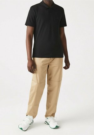 Mężczyzna w czarnej koszulce polo z krótkim rękawem, beżowych spodniach z szwem na kolanach i białych tramach z zielonymi akcentami, stojący na jednolitym tle.