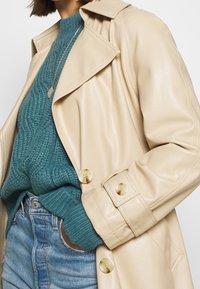 Trench coat in ecopelle beige con ampi rever, tasche laterali e bottoni oversize, indossato sopra un maglione in teal a trama e jeans di denim chiaro.