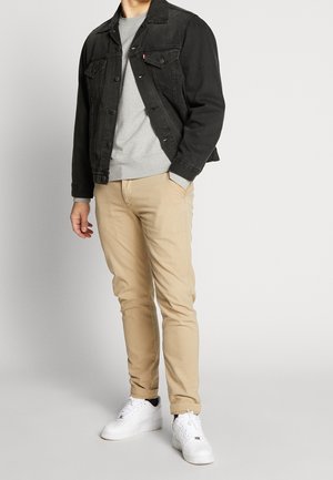 Man met zwarte spijkerjack, grijze trui, beige broek en witte sneakers, staand met één hand in de zak tegen een effen achtergrond.