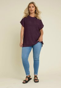 Lila texturerad topp med korta ärmar, kombinerad med ljusblå tajta jeans och svarta sandaler med spänne. Modell vänder sig framåt.