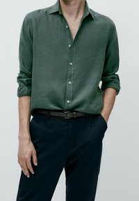 Camicia verde di lino a maniche lunghe con colletto abbottonato, maniche arrotolate e bottoni bianchi, abbinata a pantaloni blu scuro e cintura nera.