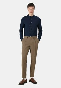 Camicia blu con colletto rialzato, abbinata a pantaloni beige su misura. Mocassini marroni completano l'outfit, con una texture liscia.