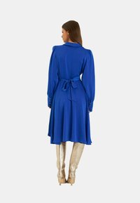 Blaues Midi-Kleid mit Puffärmel und einem Bindegürtel in der Taille. Aus Satin, luftiger Schnitt und mit geknöpften Manschetten. Getragen mit metallischen kniehohen Stiefeln.