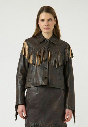 Donna che indossa una giacca in pelle marrone scuro e una gonna abbinata con dettagli frangiati marrone chiaro sul petto e sulle maniche, in piedi davanti a uno sfondo semplice.