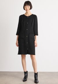 Abito in tweed nero con scollatura tonda, maniche lunghe, sei bottoni davanti e due tasche frontali. Orlo sfrangiato e tessuto texture.