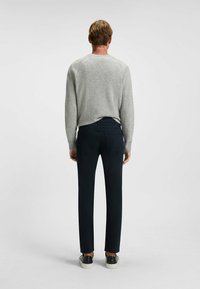 Uomo in piedi di spalle, che indossa un maglione grigio chiaro, pantaloni slim-fit scuri e sneaker nere con suole bianche, su uno sfondo semplice.