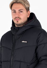 Jonge man met licht haar, gekleed in een zwart pufferjack met capuchon en het logo van "ellesse" op de borst, kijkend naar voren tegen een witte achtergrond.