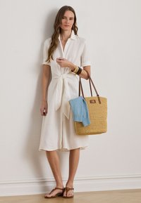 Bela lanena obleka s kratkimi rokavi in pasom za zavezovanje. Rjavi usnjeni trakovi na pleteni slamnat torbi s svetlo modrim puloverjem, ki je v notranjosti. Sandali.