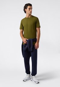 Mężczyzna w oliwkowej koszulce, granatowych spodniach dresowych, białych trampkach i granatowym rozpinanym swetrze przewiązanym w talii, stojący na jednolitym tle.