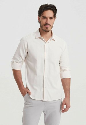 Chemise beige clair à boutons avec manches longues, dotée de poignets à revers et d'un col classique. Tissu lisse avec une texture subtile.