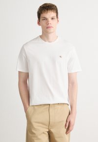 T-shirt branca de algodão com mangas curtas, decote redondo e pequeno logótipo vermelho no lado esquerdo do peito. Combinada com calças bege.