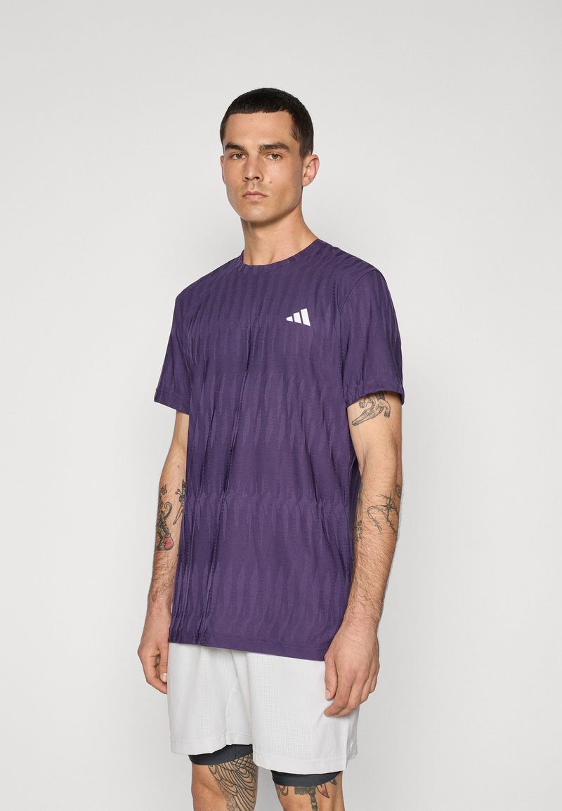 Lilla atletisk t-shirt med tekstureret mønster, rund hals, korte ærmer og hvidt logo på venstre bryst. Modellen har grå shorts på.