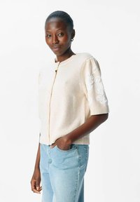 Kvinna med mörk hud som bär en krämfärgad stickad cardigan med blommönster på ärmarna och ljusblå jeans, med ena handen i fickan.